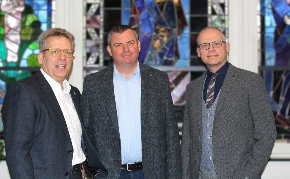 Das Direktoriun steht vor einem Mosaikfenster. Die Herren Johannes Brumm, Marcus Knittel und Dr. Michael Ludwig lächeln in die Kamera.