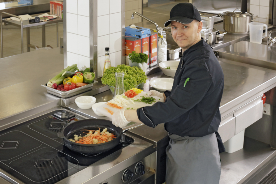 Unser Koch Herr Tollning schaut lächelnd in die Kamera, während er ein Gericht zubereitet. 