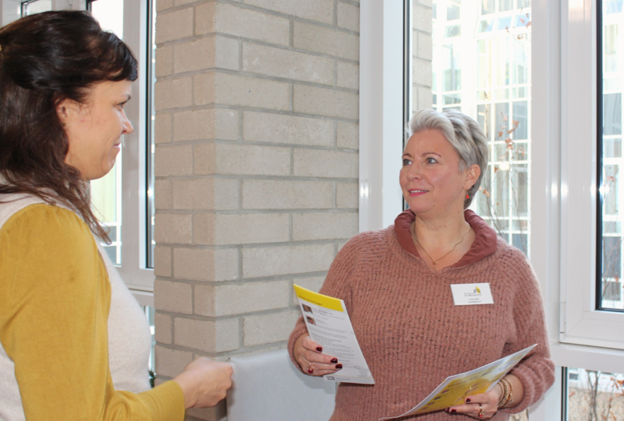Seitlich links steht eine Mitarbeiterin, die einen Flyer von einer Kollegin überreicht bekommt.