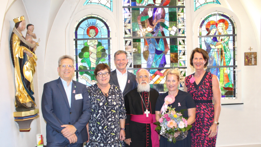 Auf dem Foto erkennen Sie von links nach rechts Johannes Brumm, Geschäftsführer, Petra Grimm-Benne, Gesundheitsministerin; Dr. med. Wolfgang Hartmann, Chefarzt der Frauenheilkunde und Geburtshilfe; Bischof Dr. Gerhard Feige; Dr. med. Kristina Freese, Leidende Oberärztin des zertifizierten Brustzentrums und Dr. med. Anke Kleine-Tebbe, Chefärztin des zertifizierten Brustzentrums Köpenick der DRK Kliniken Berlin. Die Gruppe steht vor einem bunten Mosaikfenster.